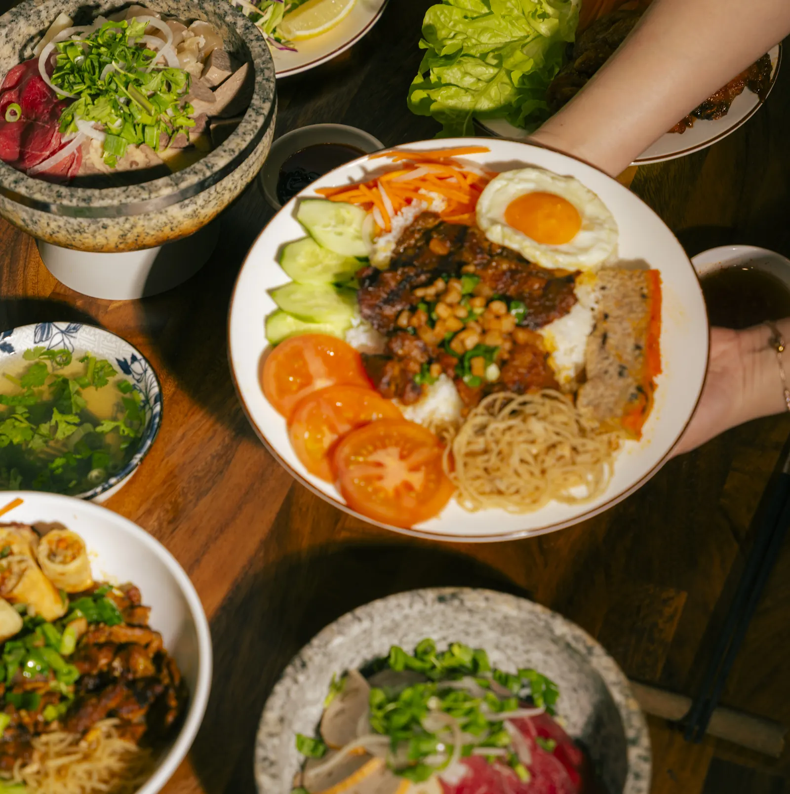 A full table of Vietnamese dishes being served at Bà Năm Restaurant, Adelaide — cơm tấm, stone-bowl phở and fresh herbs