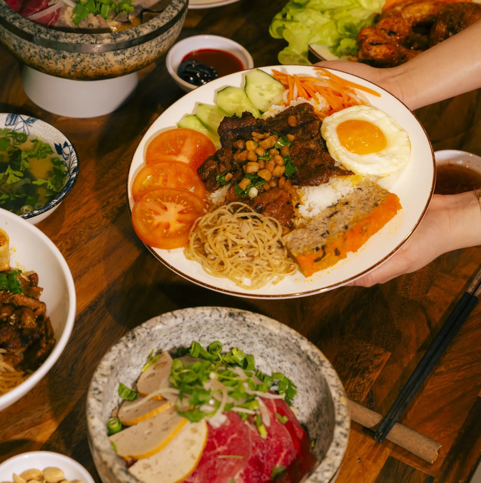 Overhead view of a shared Vietnamese lunch at Bà Năm Adelaide — hands holding a broken-rice plate with grilled pork, egg, chả, tomato and cucumber, a stone bowl of phở with paper-thin raw red-coin beef in the foreground, and more bowls across a wooden table.