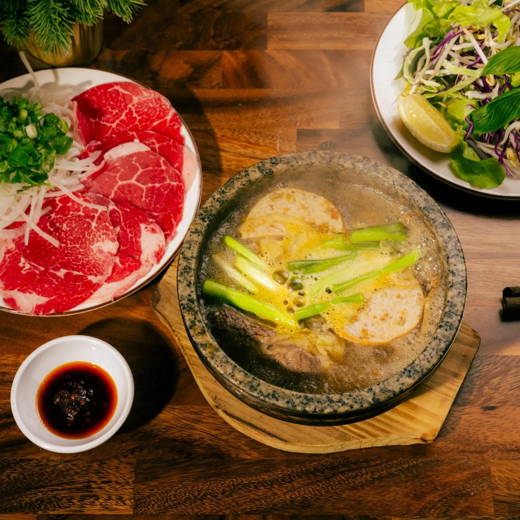 Stone bowl of bún bò Huế with amber lemongrass-tinged broth, sliced Vietnamese pork sausage, spring onion and peppercorns, beside a plate of thick rare beef slices, shredded cabbage and carrot salad, basil and lemon.