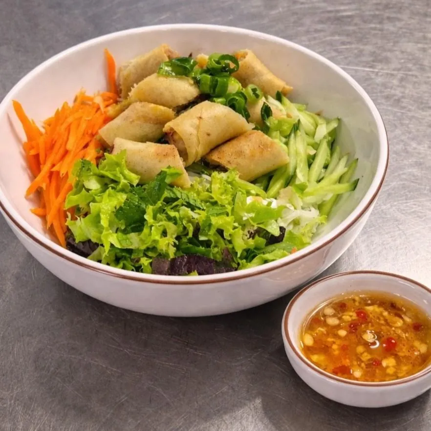 Bún Chả Giò at Bà Năm Adelaide — white ceramic bowl of rice vermicelli topped with whole golden fried spring rolls and spring onion, shredded carrot on the left, cucumber sticks on the right, lettuce and mint at the front, with a small side bowl of nước chấm with chilli and garlic.