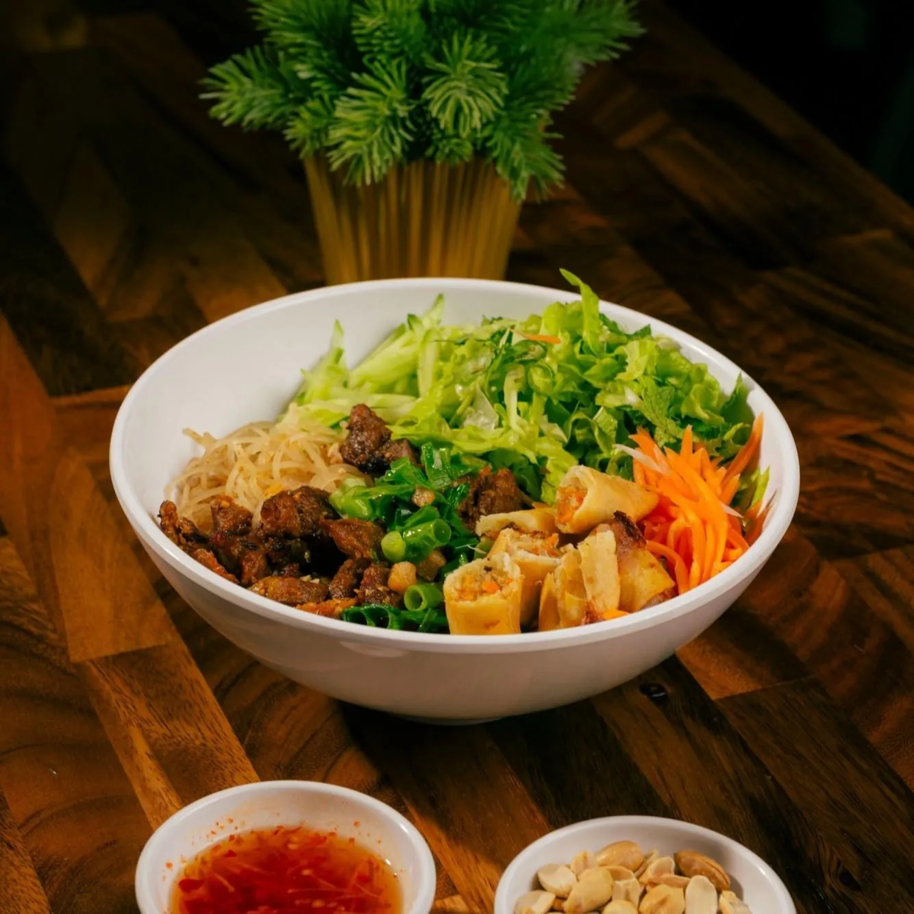 Bún Đặc Biệt at Bà Năm Adelaide: white ceramic bowl of rice vermicelli topped with caramelised grilled pork pieces, cut golden fried spring rolls, shredded carrot, cucumber sticks, shredded lettuce, mint and chopped spring onion, with side ramekins of red chilli nước chấm and peeled peanuts on a wooden table.