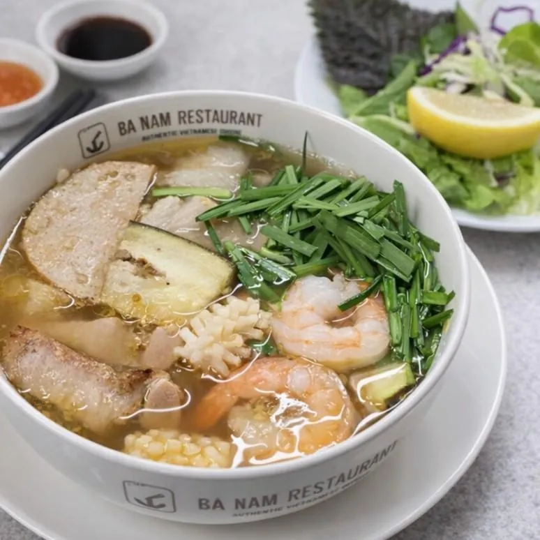 Bún Mắm at Bà Năm Restaurant Adelaide — white branded bowl of Mekong-style fermented fish noodle soup with amber broth, thick rice vermicelli, prawns, squid, pork slices, chả lụa and eggplant, topped with snipped chives, served with fresh herbs, lettuce and lemon wedge.