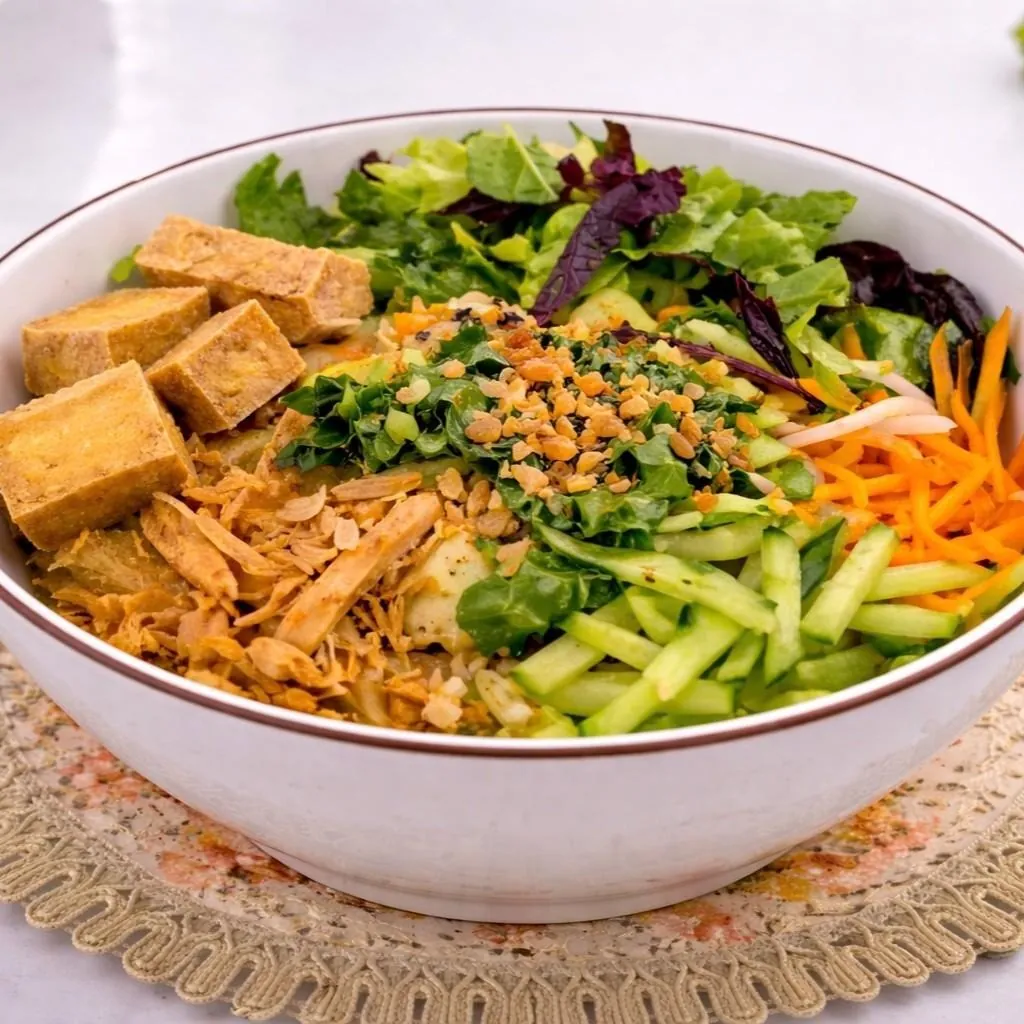 Bún Tàu Hủ Chay at Bà Năm Adelaide — white ceramic bowl with a thin brown rim on a lace doily, holding golden-brown fried tofu cubes on the left, shredded golden crispy bean curd at lower centre, mixed lettuce and purple leaf at the top, shredded carrot and bean sprouts on the right, julienned cucumber sticks at front right, and chopped green herbs with crushed peanut and fried shallot garnish in the centre.