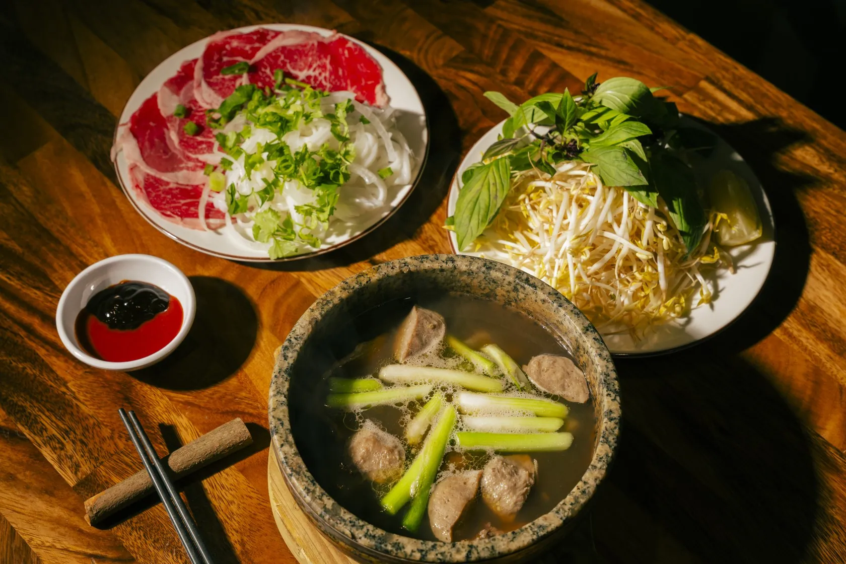 A steaming stone bowl of long-simmered broth with herbs and rare beef sides at Bà Năm Restaurant, Adelaide — the same unhurried kitchen behind our bò kho