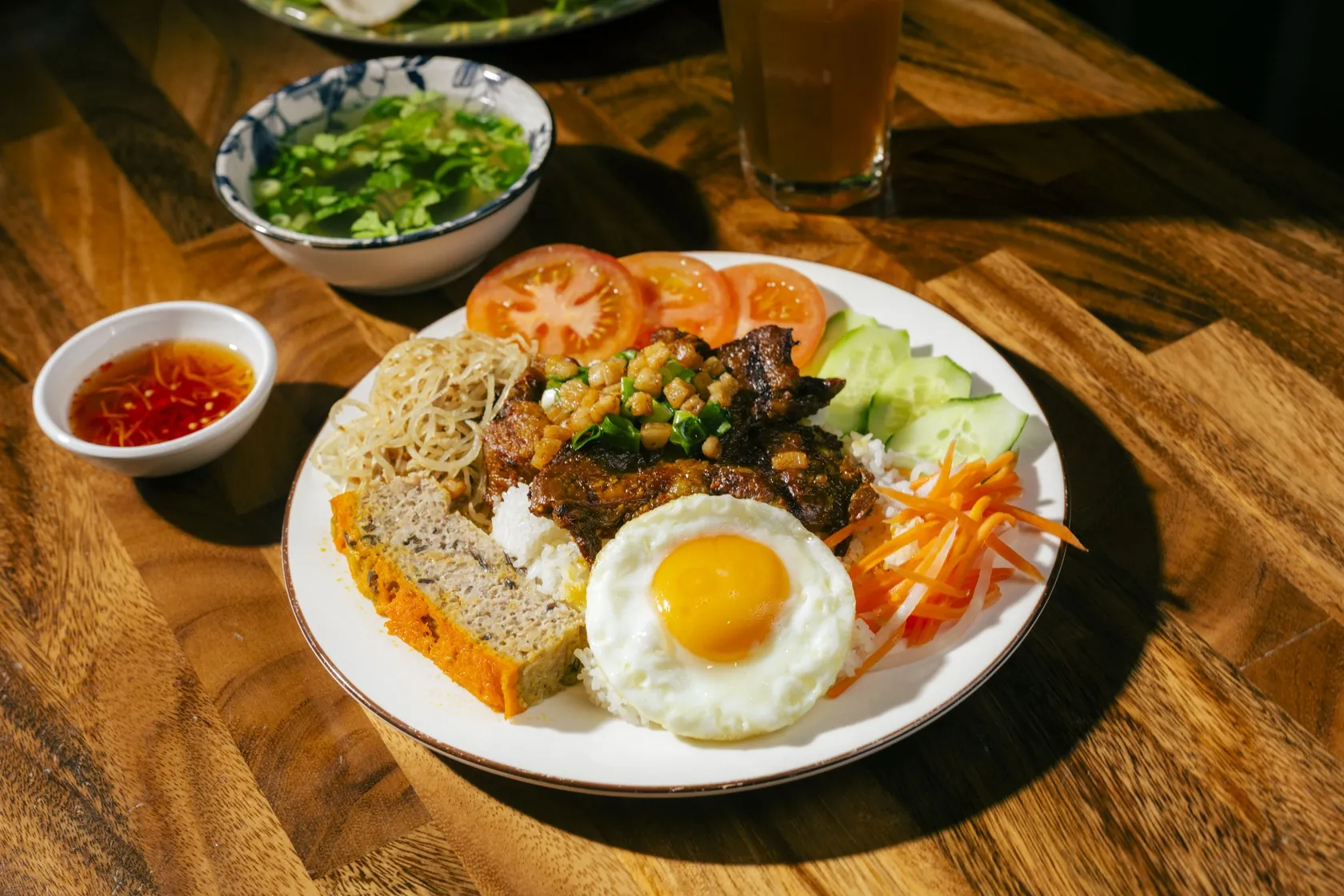 Overhead view of cơm tấm at Bà Năm Adelaide: broken rice with grilled pork, scallion and fried shallot, sunny-side fried egg, chả, pickled carrot and daikon, tomato and cucumber, with chilli nước chấm and a bowl of clear canh soup on a timber table