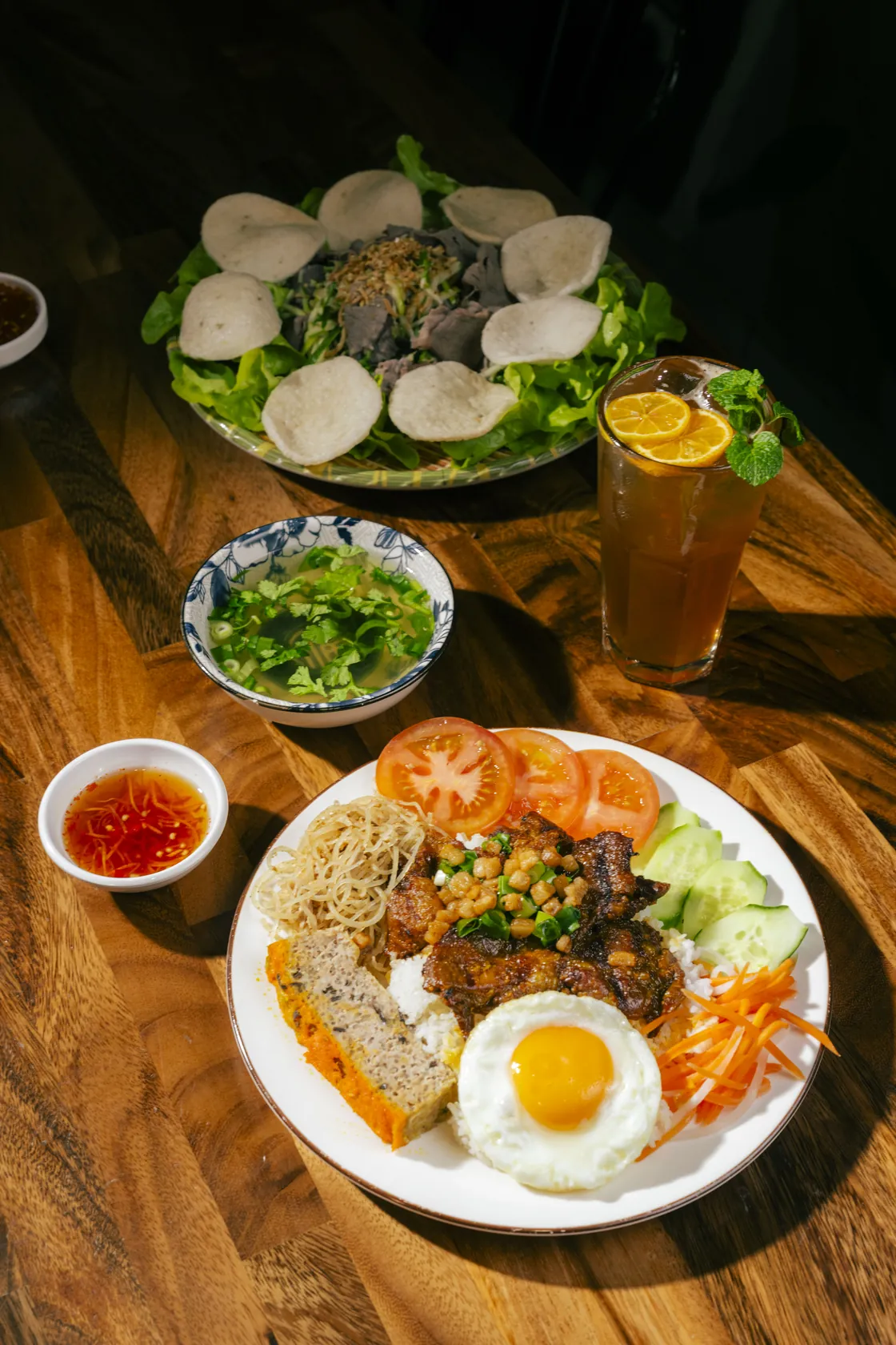 Cơm tấm with fried egg and grilled pork beside gỏi bò tái chanh with prawn crackers and iced tea on a timber table at Bà Năm