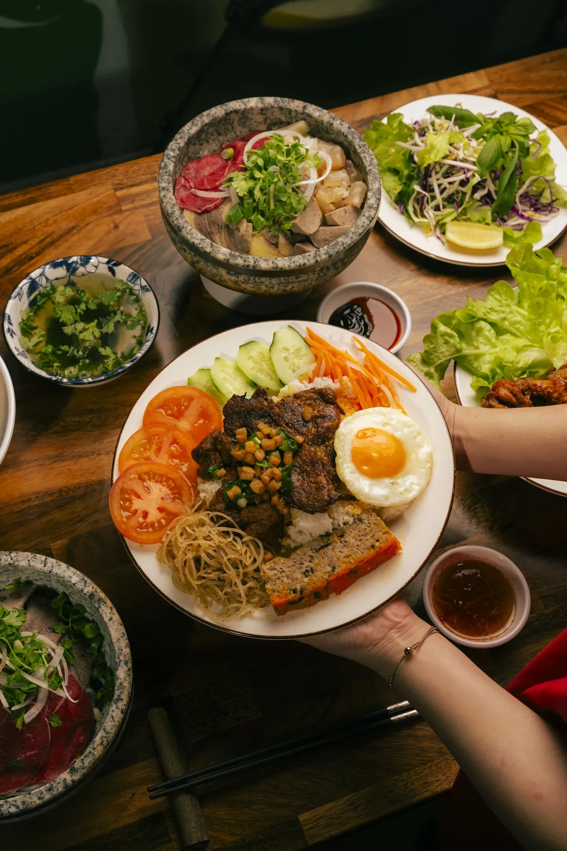 Hands presenting a cơm tấm plate with grilled pork, fried egg, chả, pickles, tomato, cucumber and broken rice, with Phở Thố Đá stone bowls and a gỏi bò tái chanh salad on the table at Bà Năm, Adelaide
