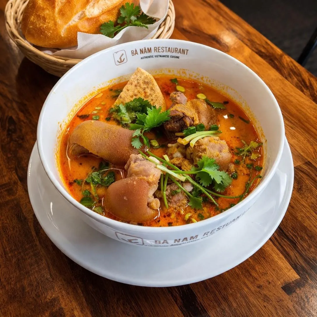Bà Năm branded white bowl of Gia Cay, orange-red turmeric and galangal pork hock stew with skin-on hock pieces, coriander and spring onion, with a bread roll in a basket behind, at Bà Năm Restaurant Adelaide.