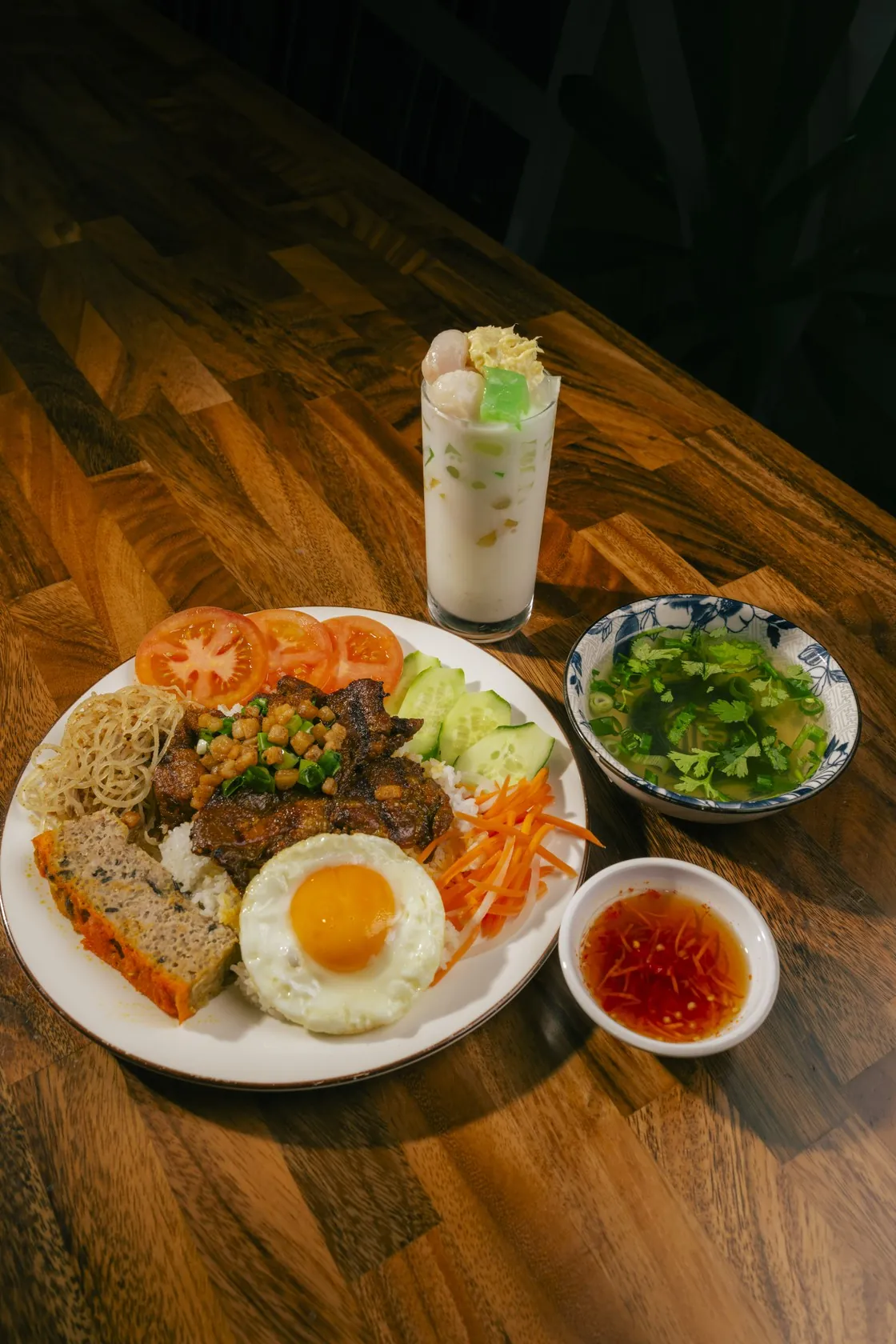 Cơm tấm plate with grilled pork, egg, chả, pickles, clear soup, nước chấm and a tall iced chè drink on a wooden table at Bà Năm, Chinatown Adelaide