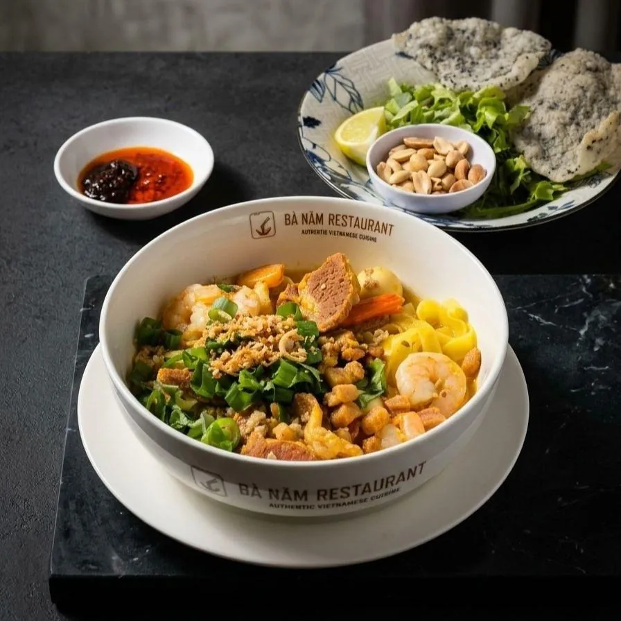 Bowl of mì quảng at Bà Năm Adelaide with wide turmeric-gold noodles, prawns, pork, fish cake, crushed peanuts and fried shallots, served with rice crackers and lettuce on the side