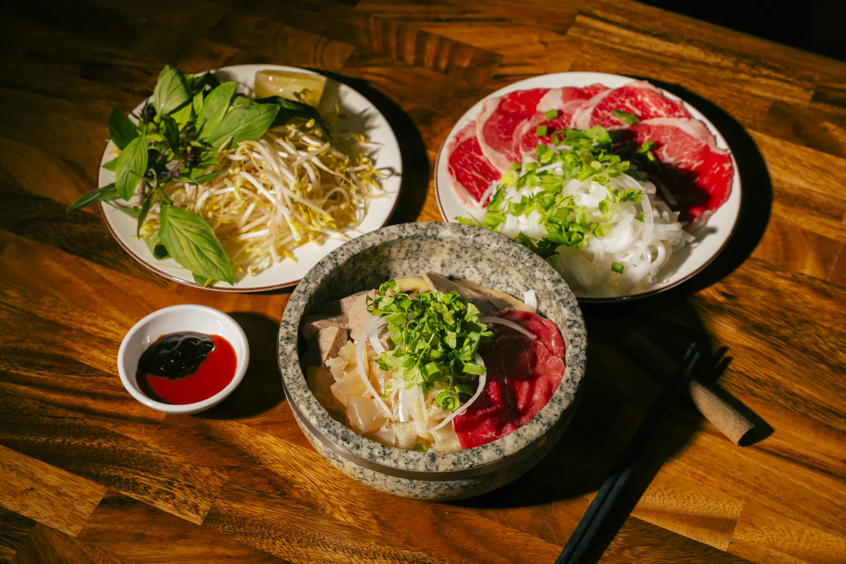 Stone bowl phở at Bà Năm Adelaide with a side plate of fresh herbs and bean sprouts and a plate of paper-thin raw beef ready to cook in the broth