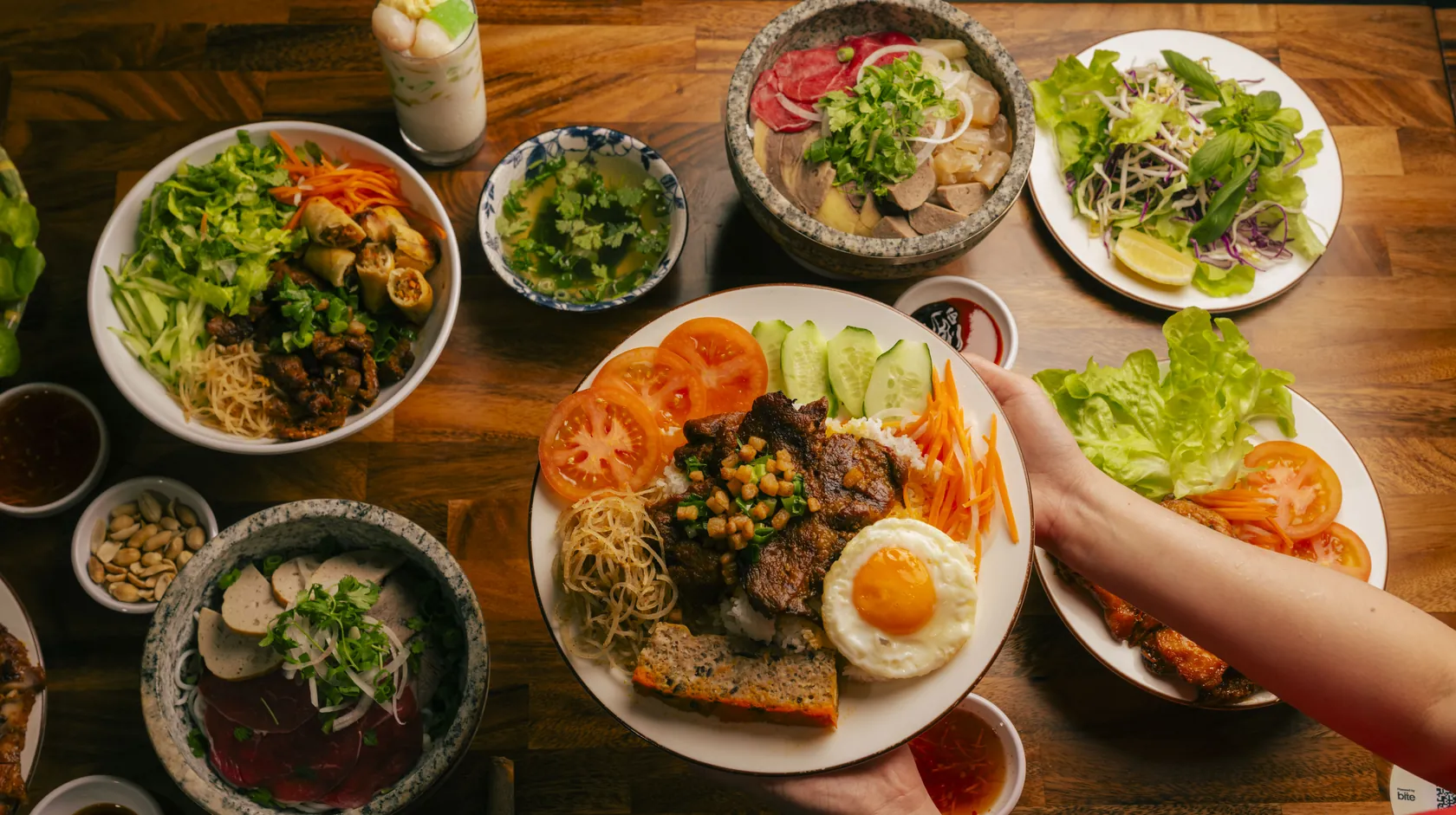 Overhead share table at Bà Năm, 65 Grote Street Adelaide — hands holding cơm tấm with grilled pork, fried egg and chả on broken rice, a stone bowl phở with rare beef, tendon and tripe, bún vermicelli with grilled meat and spring rolls, gỏi, fresh herbs and bean sprouts, and a tall glass of chè thái