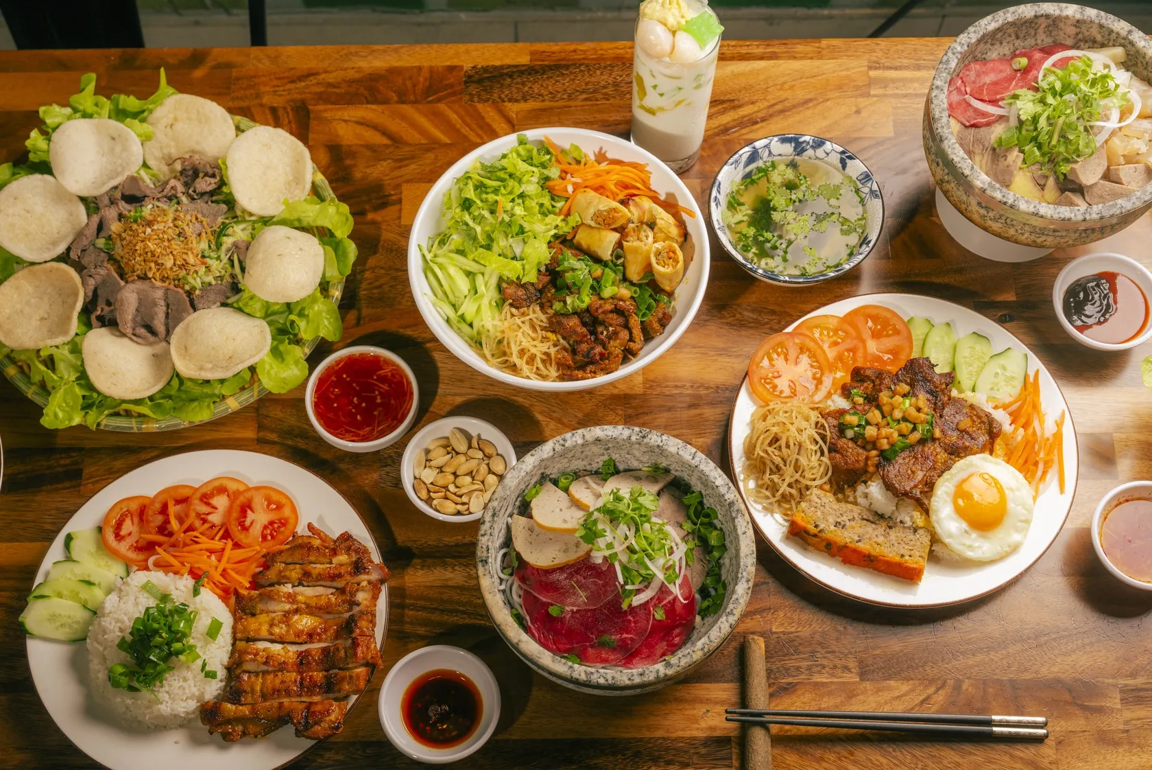 Overhead Vietnamese share table on timber at Bà Năm Adelaide — gỏi bò tái chanh with prawn crackers, bún vermicelli with grilled meat and spring rolls, two stone-bowl phở with raw red-coin beef, grilled pork rice, cơm tấm with egg and chả, and a chè thái