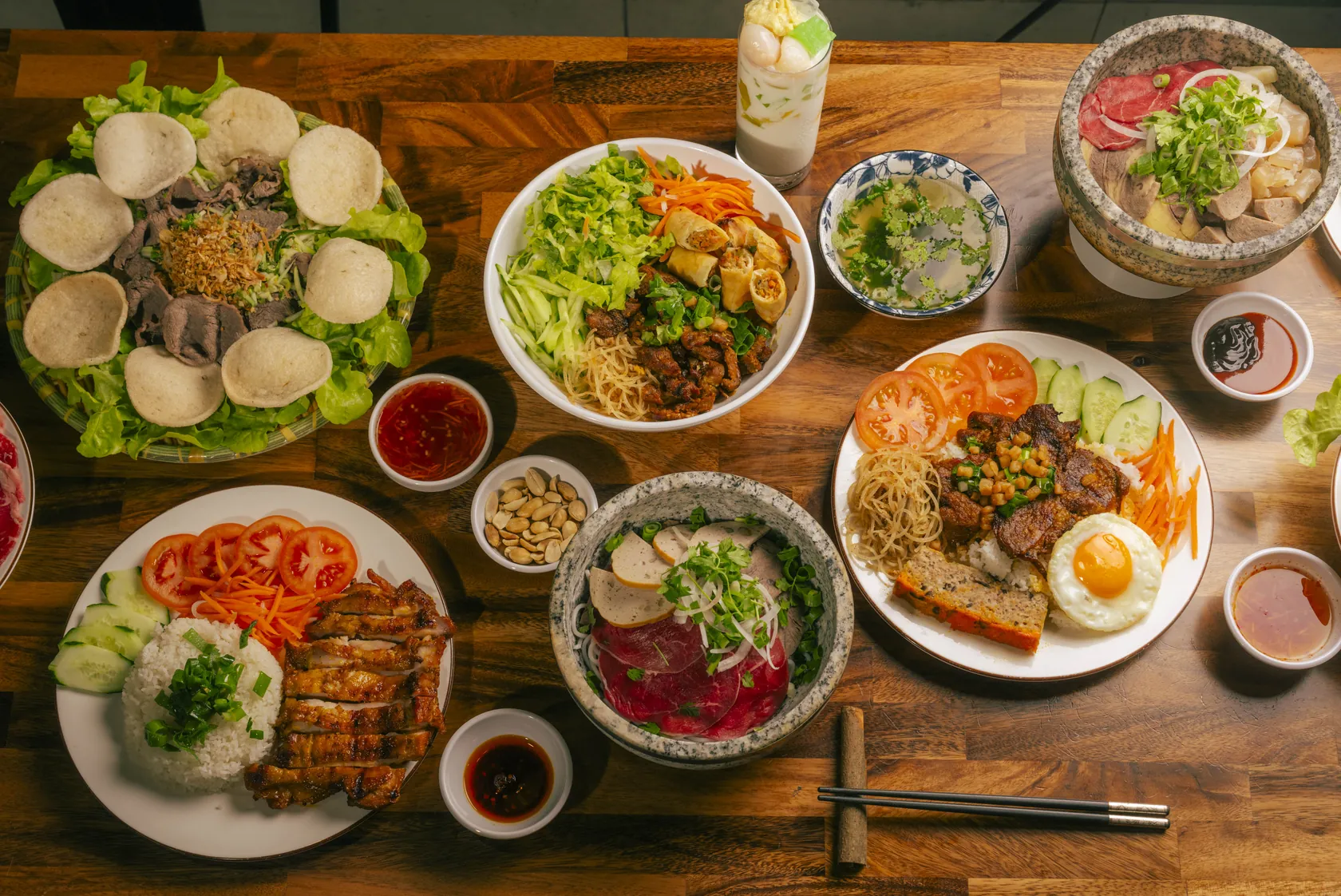 Overhead share table at Bà Năm Adelaide with two granite stone bowls of phở, cơm tấm with grilled pork and fried egg, grilled chicken rice, gỏi bò tái chanh with prawn crackers, bún vermicelli with spring rolls, chè thái, sauces and chopsticks
