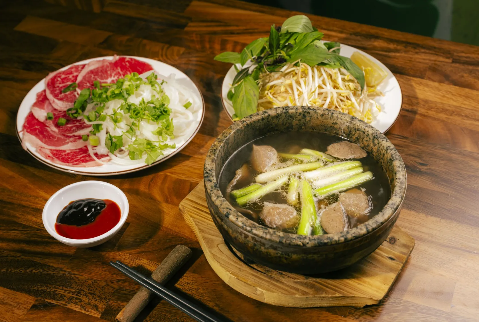 Phở Thố Đá at Bà Năm, 65 Grote Street Adelaide — dark granite stone bowl of steaming beef broth with meatballs and long spring onion stalks, side plate of paper-thin red-coin rare beef with onion, coriander and spring onion, herb plate with bean sprouts, Thai basil and lemon, soy dip and chopsticks on timber