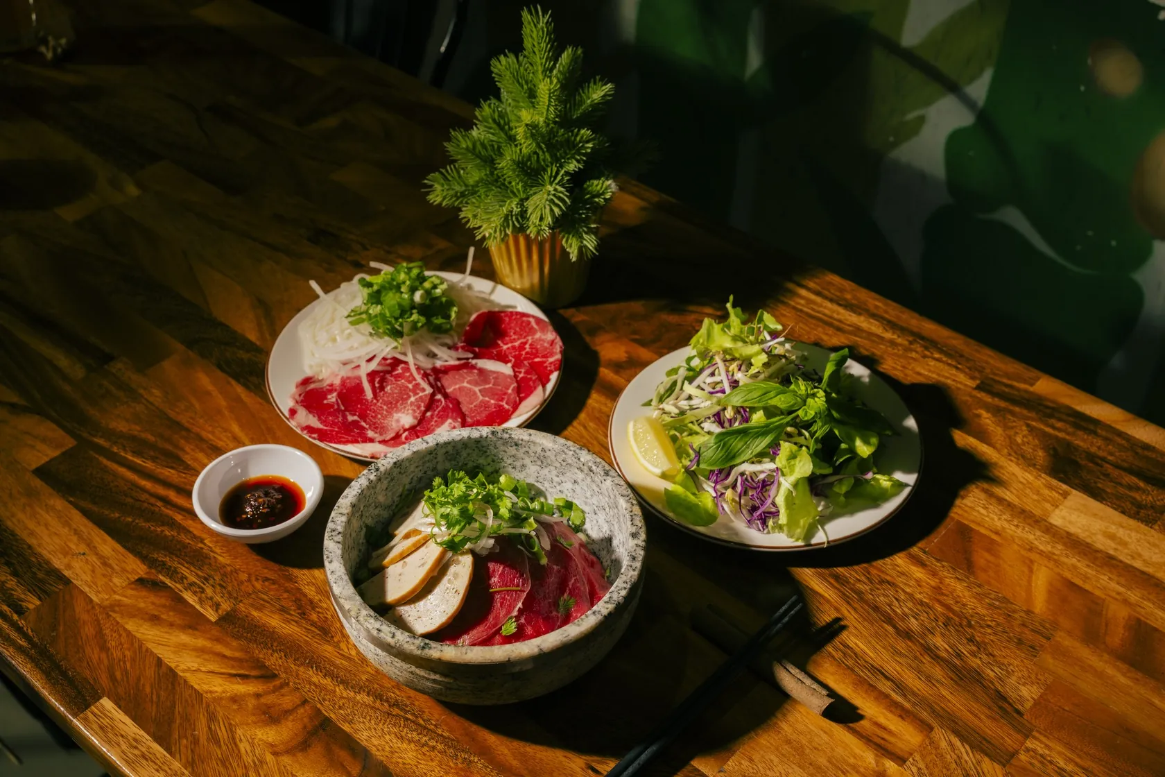 Phở Thố Đá at Bà Năm, 65 Grote Street Adelaide — grey stone bowl with rare beef and fish cake slices under herbs, side plate of paper-thin red-coin rare beef with onion and spring onion, herb and cabbage salad with lemon wedge, small soy dip and chopsticks on a timber table
