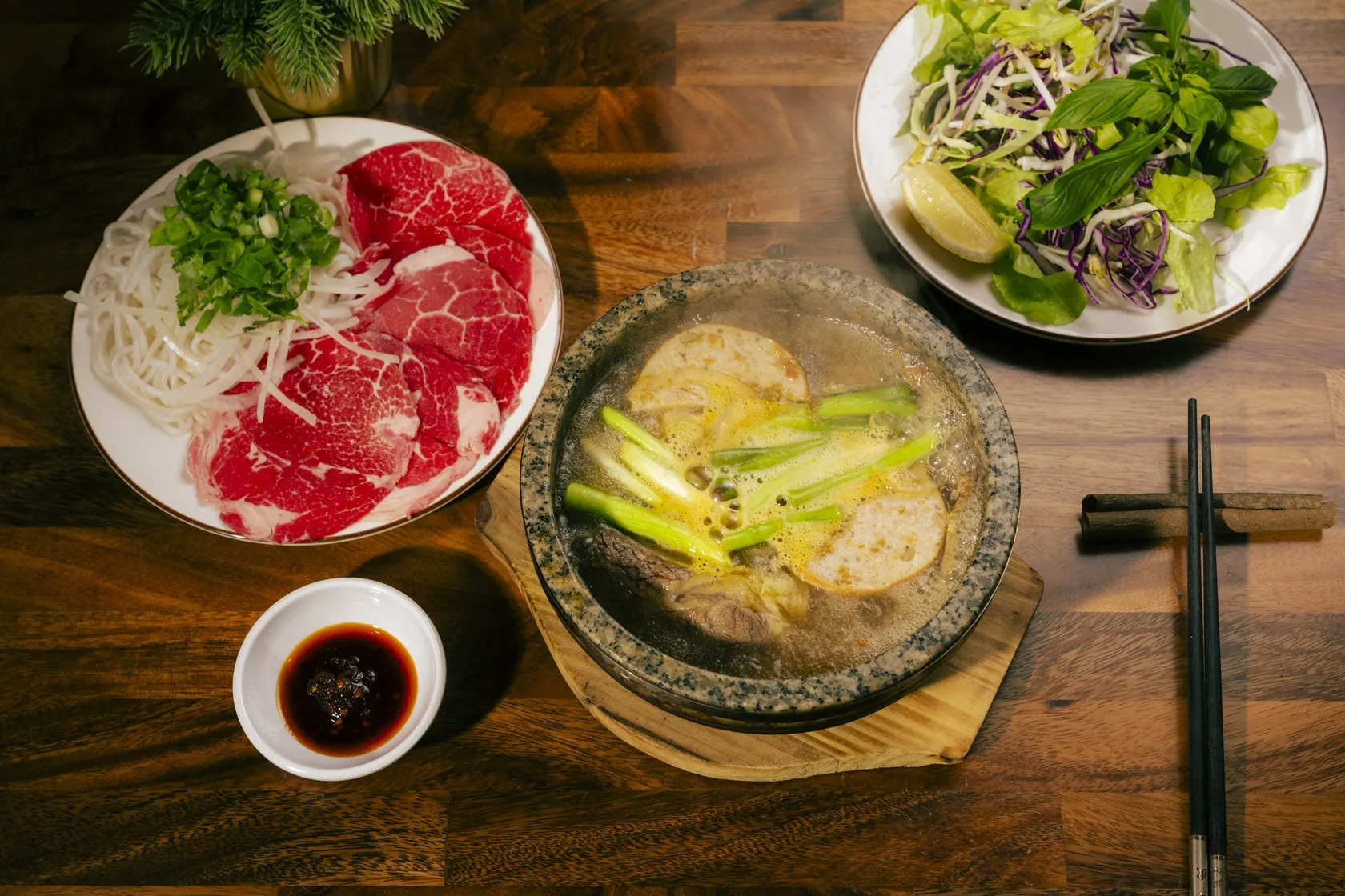 Overhead Phở Thố Đá stone-bowl set at Bà Năm Adelaide with raw red-coin rare beef, fresh herbs and chilli sauce on a timber table