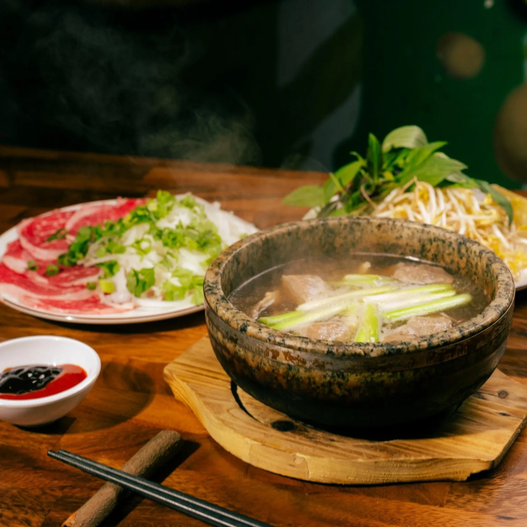 Steaming stone bowl of clear phở broth with spring onion and cooked beef on a wooden trivet at Bà Năm, with a side plate of paper-thin raw rare beef, rice noodles, fresh herbs, bean sprouts and hoisin-chilli sauces.
