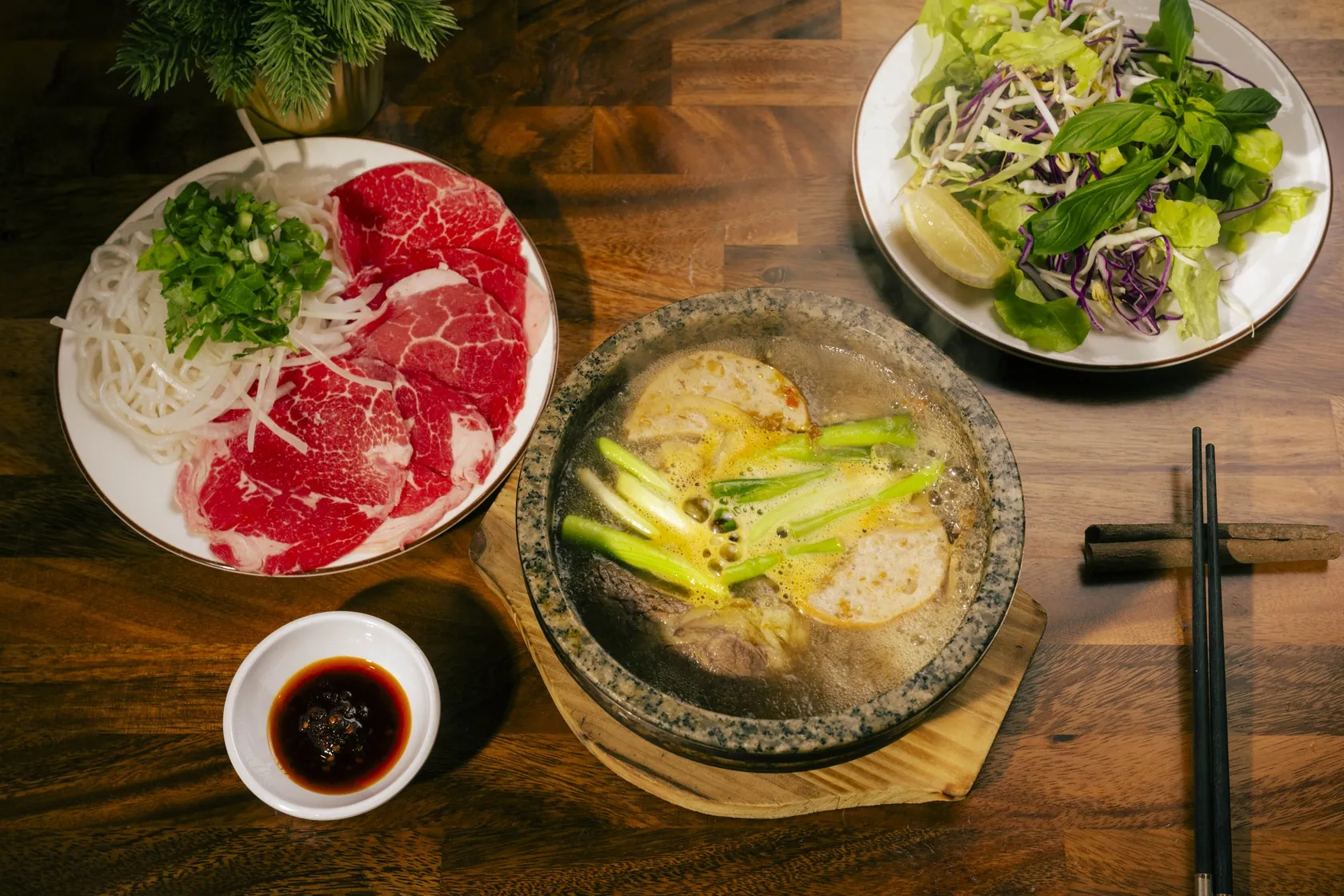Overhead view of Bà Năm's stone bowl phở set on a timber table: steaming stone bowl with green onion and fish cake, side plate of raw red-coin beef with rice noodles, herb plate with lemon and chilli sauce