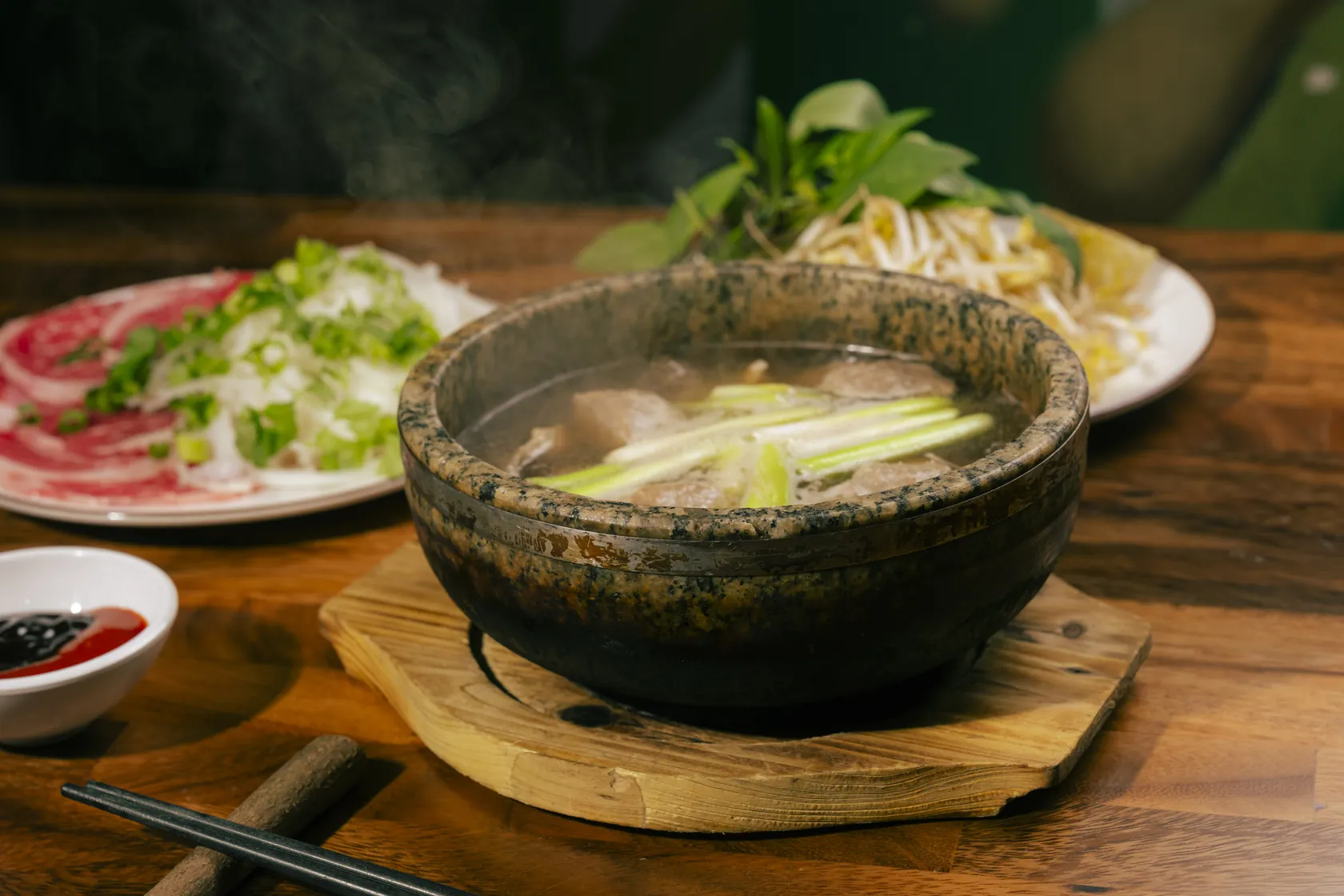 Steaming granite stone bowl of Phở Thố Đá with cooked beef and long spring onion in clear broth on a wooden trivet, beside a plate of paper-thin red-coin rare beef with white onion and spring onion, a plate of fresh herbs, bean sprouts and lime, a small chilli sauce dish and black chopsticks on a wooden table at Bà Năm, Adelaide Chinatown