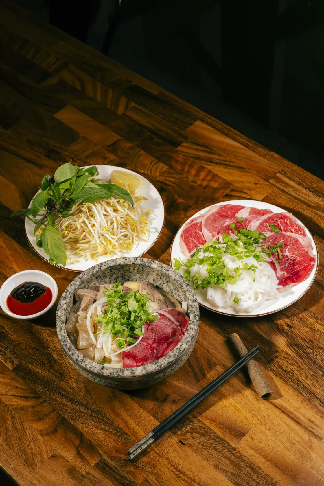 Overhead view of Phở Thố Đá stone bowl set at Bà Năm Adelaide: grey granite stone bowl with sliced rare beef, tendon and spring onion, side plate of raw red-coin beef with onion and herbs, bean sprouts and Thai basil plate, chilli sauce and chopsticks on timber table