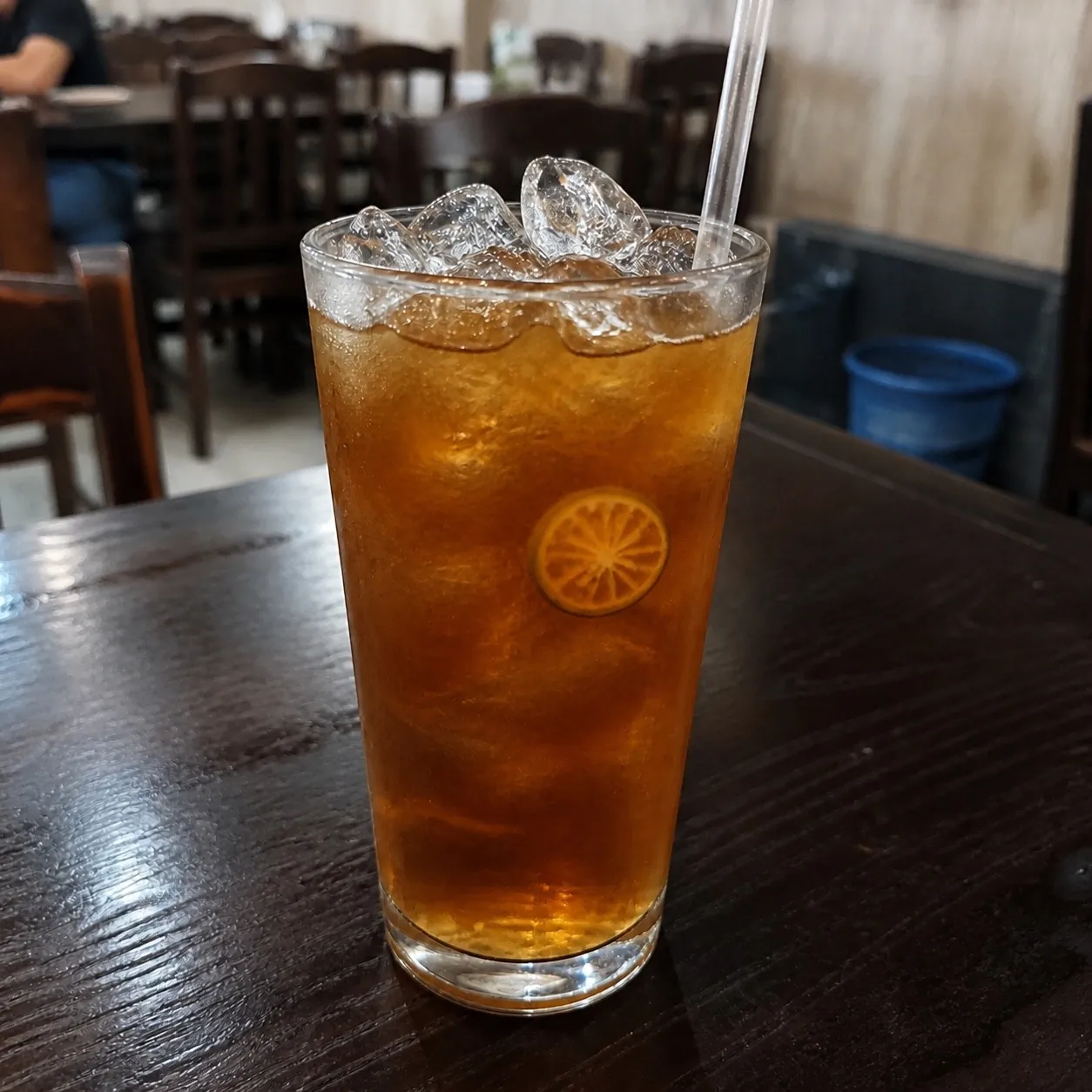 Tall clear glass of amber-brown Trà Tắc kumquat iced tea packed with ice, a dried citrus wheel in the liquid, on a dark wooden table at Bà Năm, Adelaide
